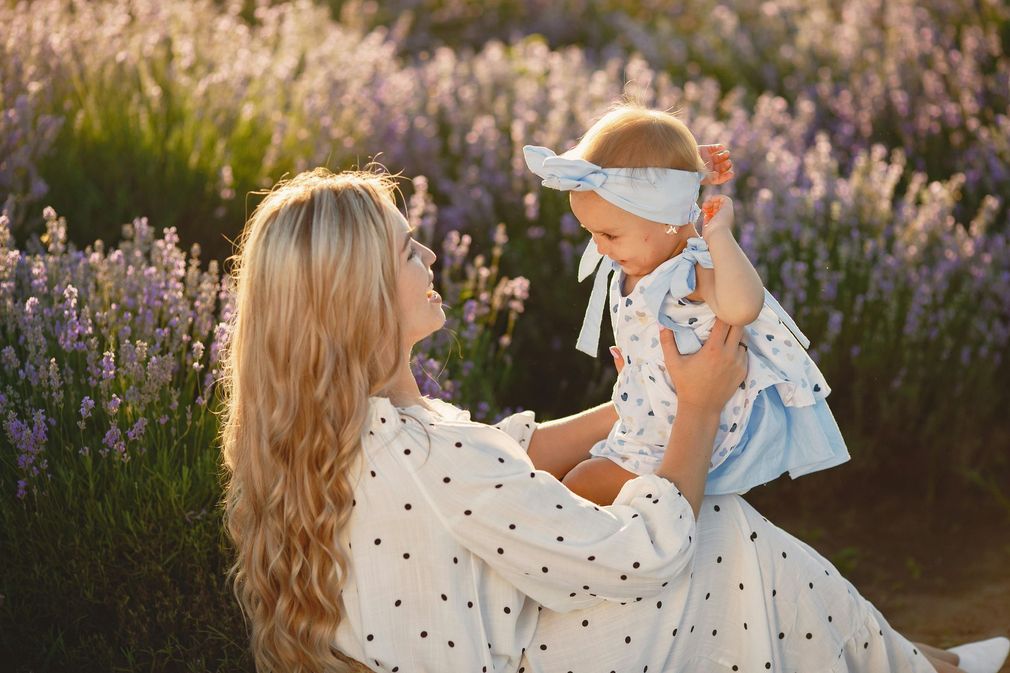 Woman throws a baby into the air, laughing, in front of a lilac bush.