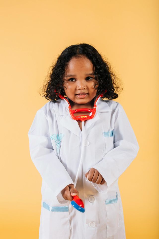 Girl wearing a doctor's coat and stethoscope, looking curious, on a yellow background.