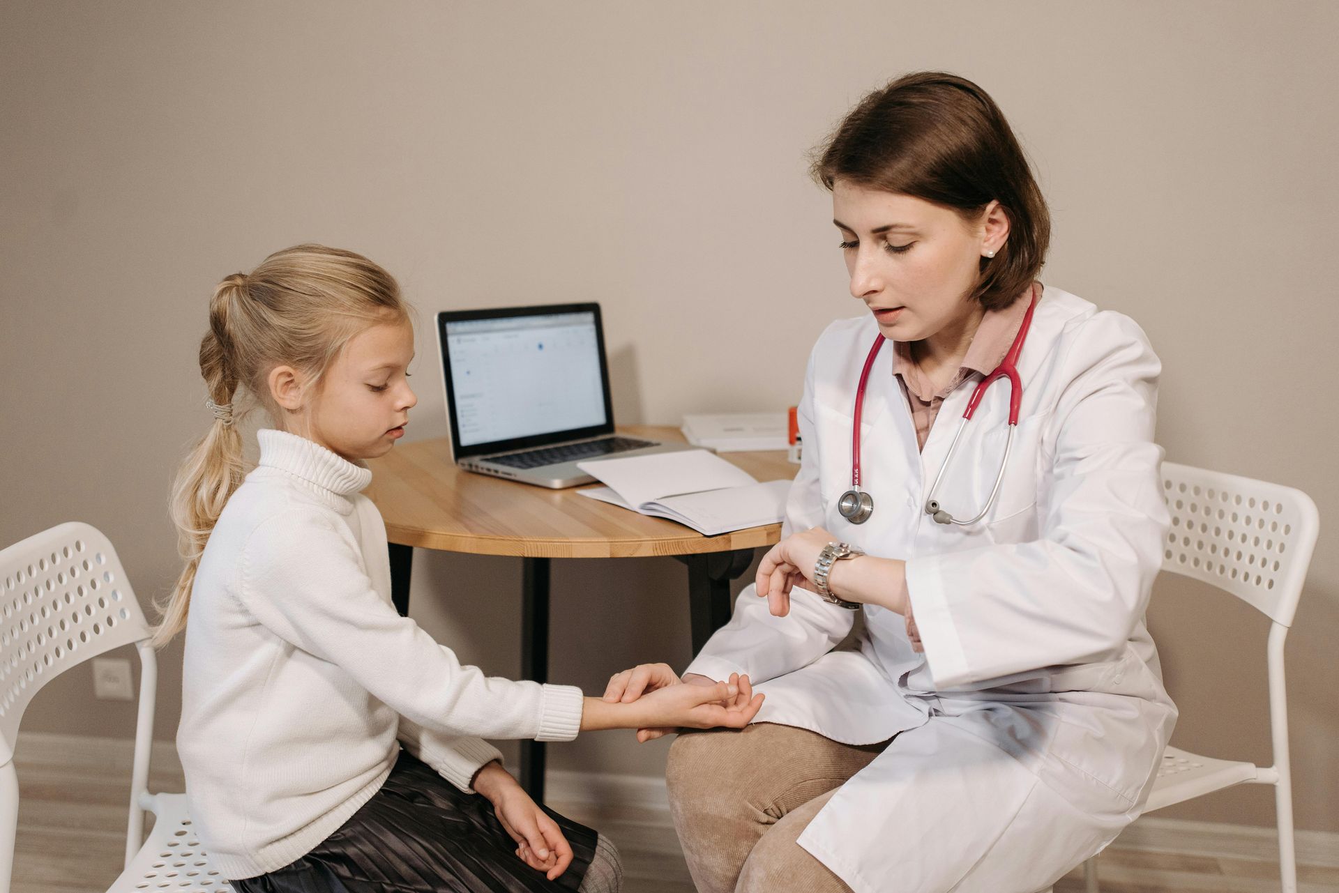 Doctor checking a child's pulse in a medical office. The doctor has a stethoscope and the child is wearing a white sweater.