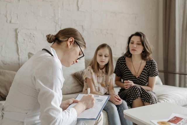 A healthcare provider writes on a clipboard, meeting with a mother and child seated on a couch.
