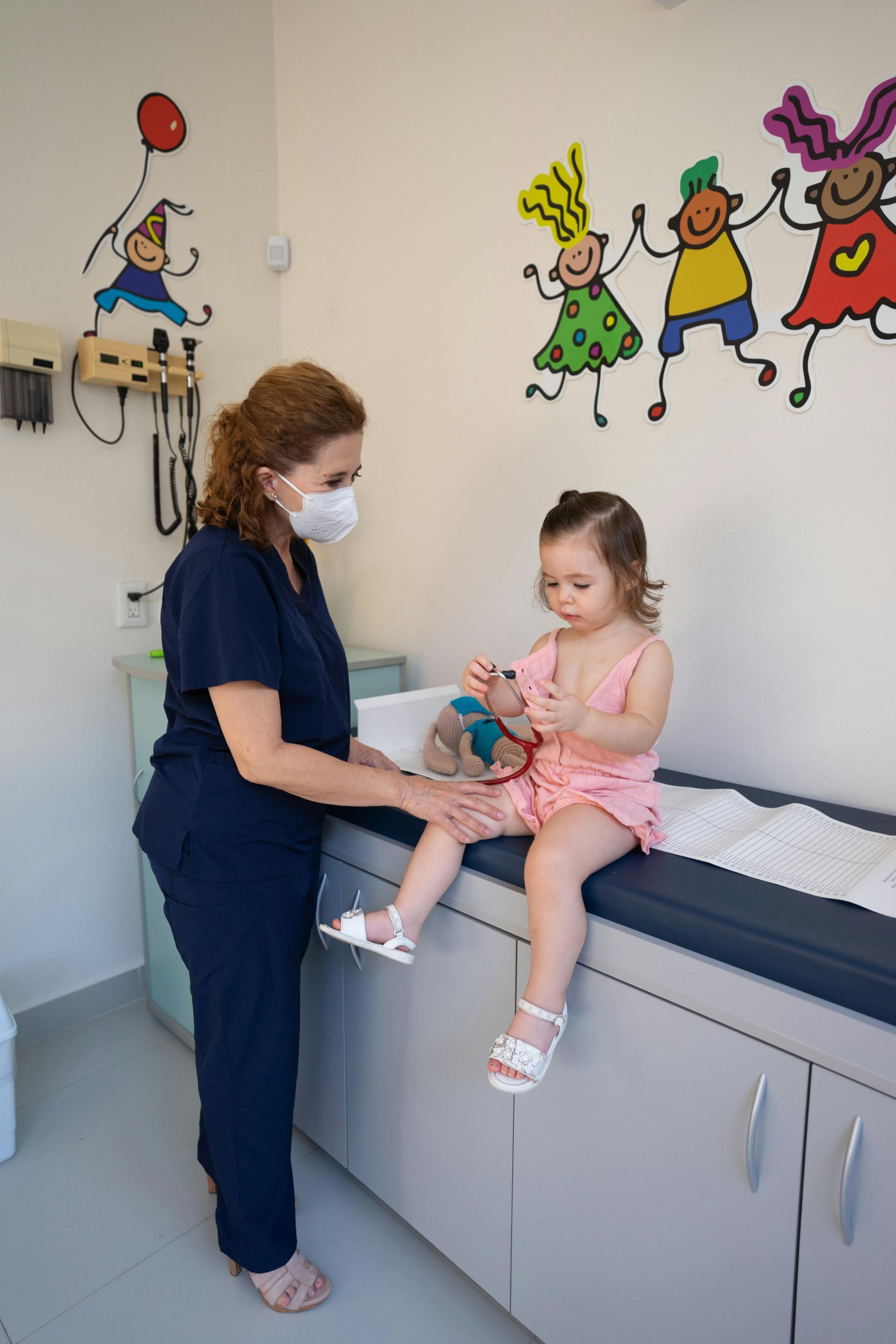 Nurse in mask examines a young child's leg on an exam table in a brightly decorated clinic.