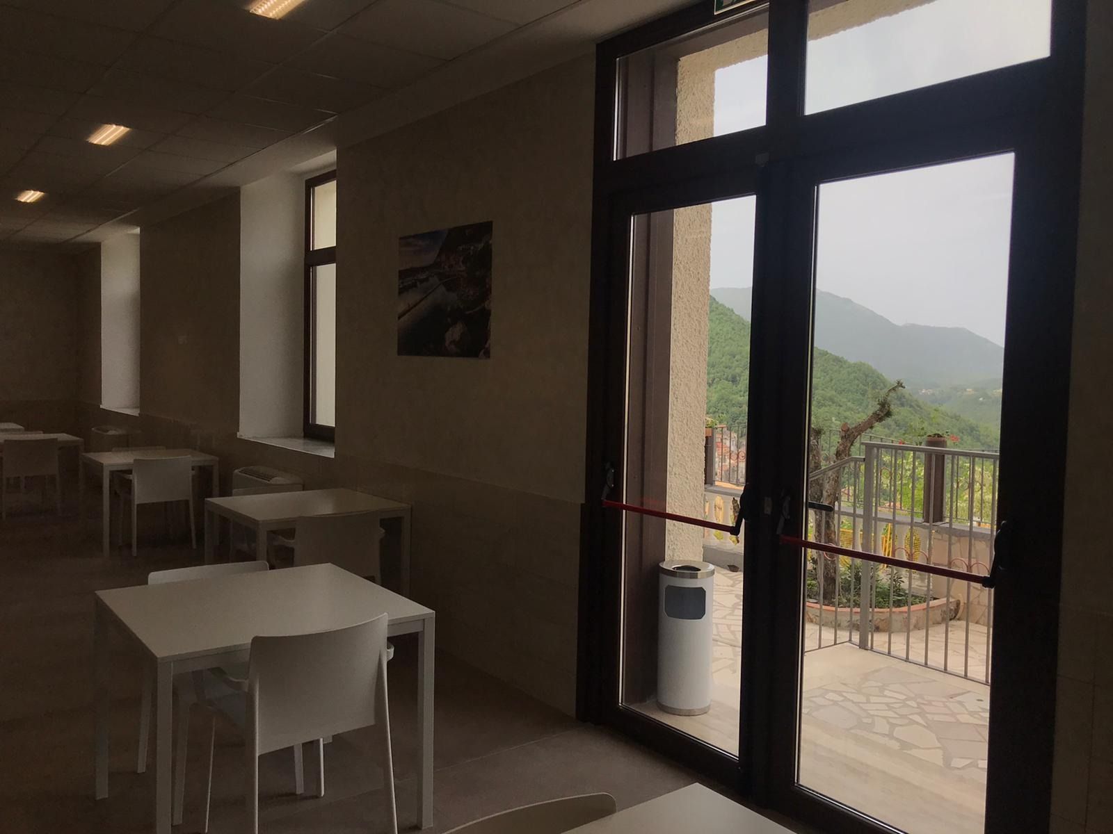 Sala interna con tavoli, sedie e ampia finestra con vista su un paesaggio collinare e verdeggiante.