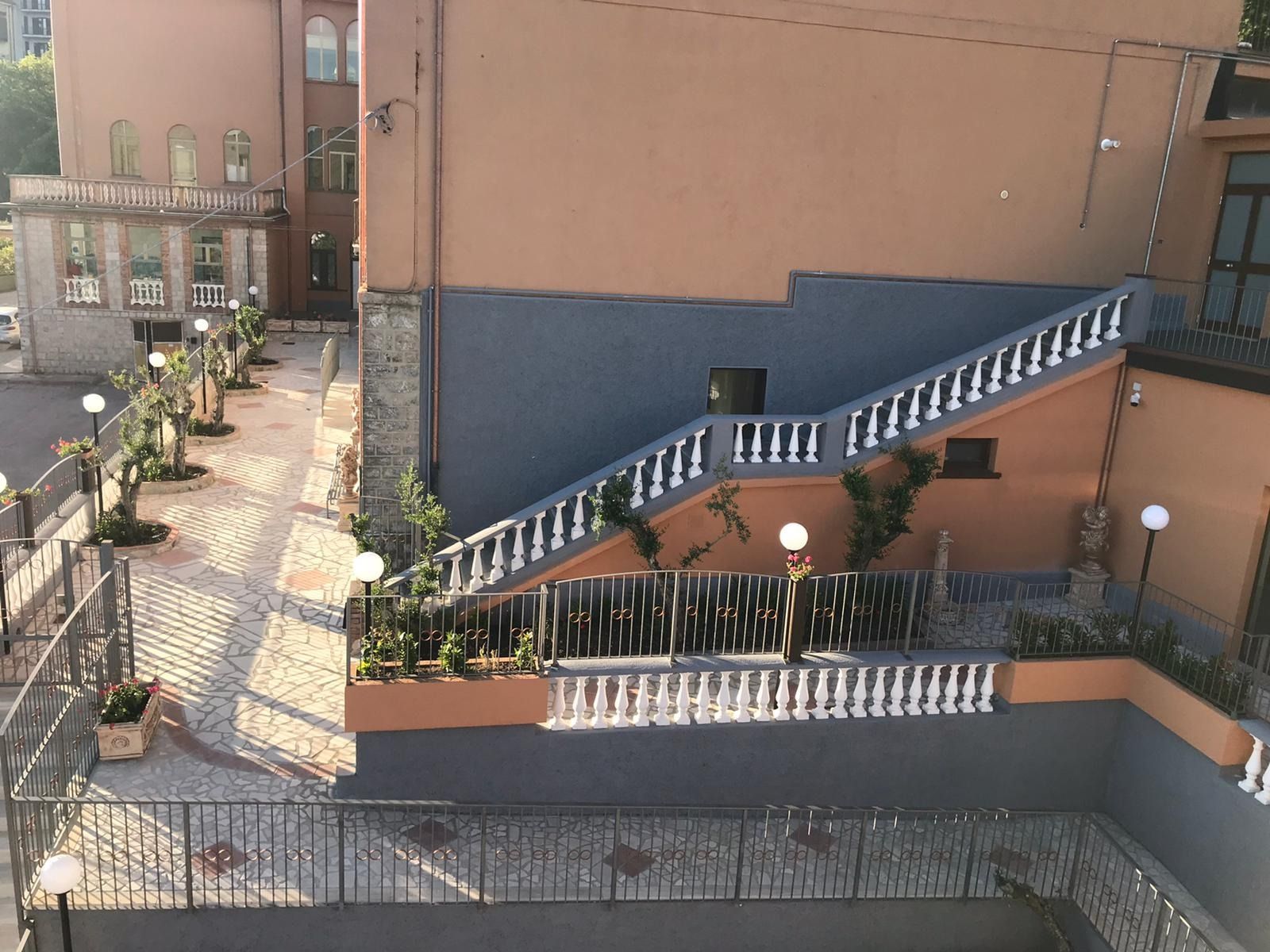 Scala esterna in pietra con balaustre bianche, che sale da un cortile pavimentato accanto a un edificio color pesca.