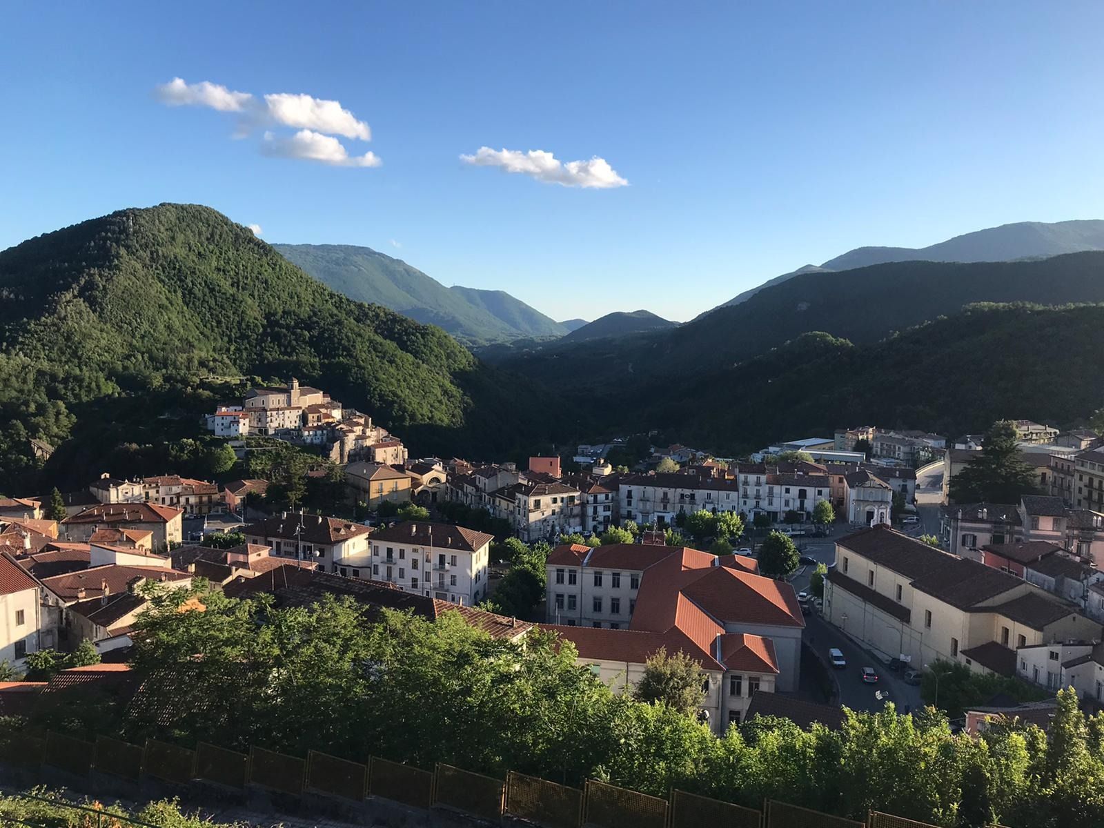 Città adagiata in una valle con montagne sotto un cielo azzurro, con edifici dai tetti di tegole rosse.