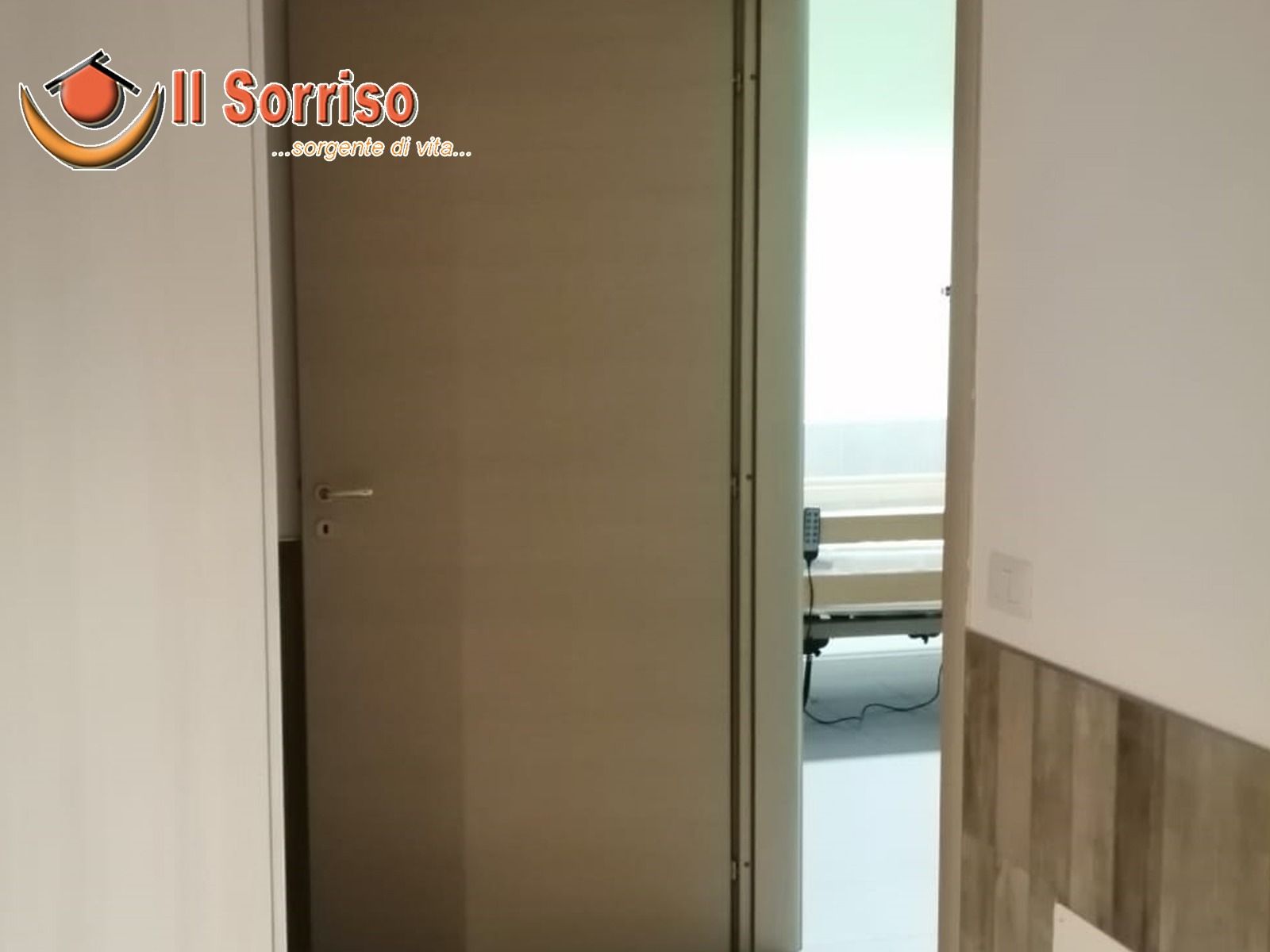 Porta di una stanza d'ospedale con un letto visibile. Porta beige, pareti bianche e illuminazione intensa.