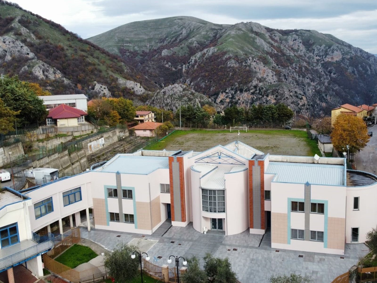 Edificio scolastico con montagne sullo sfondo; esterno beige e blu, prato verde, alberi e piccole case.