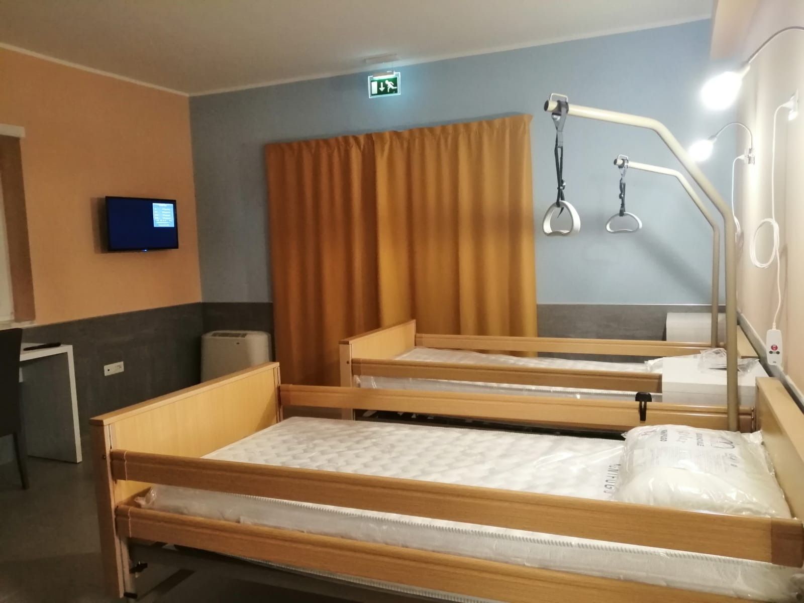 Due letti d'ospedale in una stanza con attrezzature mediche, una piccola TV e tende gialle.