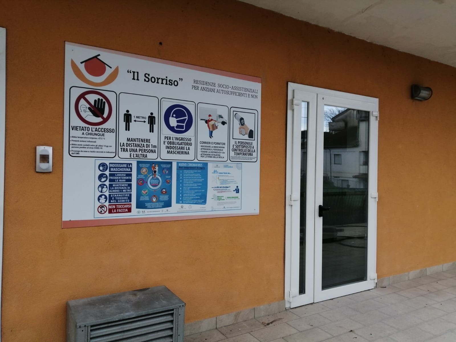 Edificio arancione con porta in vetro con cornice bianca e un cartello con le istruzioni di sicurezza; sotto c'è un piccolo riquadro grigio.