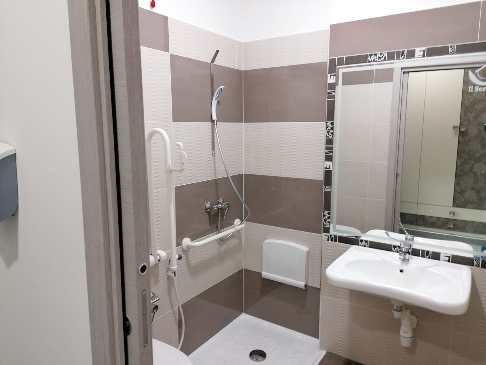 Bagno con doccia accessibile, lavandino bianco e corrimano. Pareti piastrellate marroni e beige e specchio.
