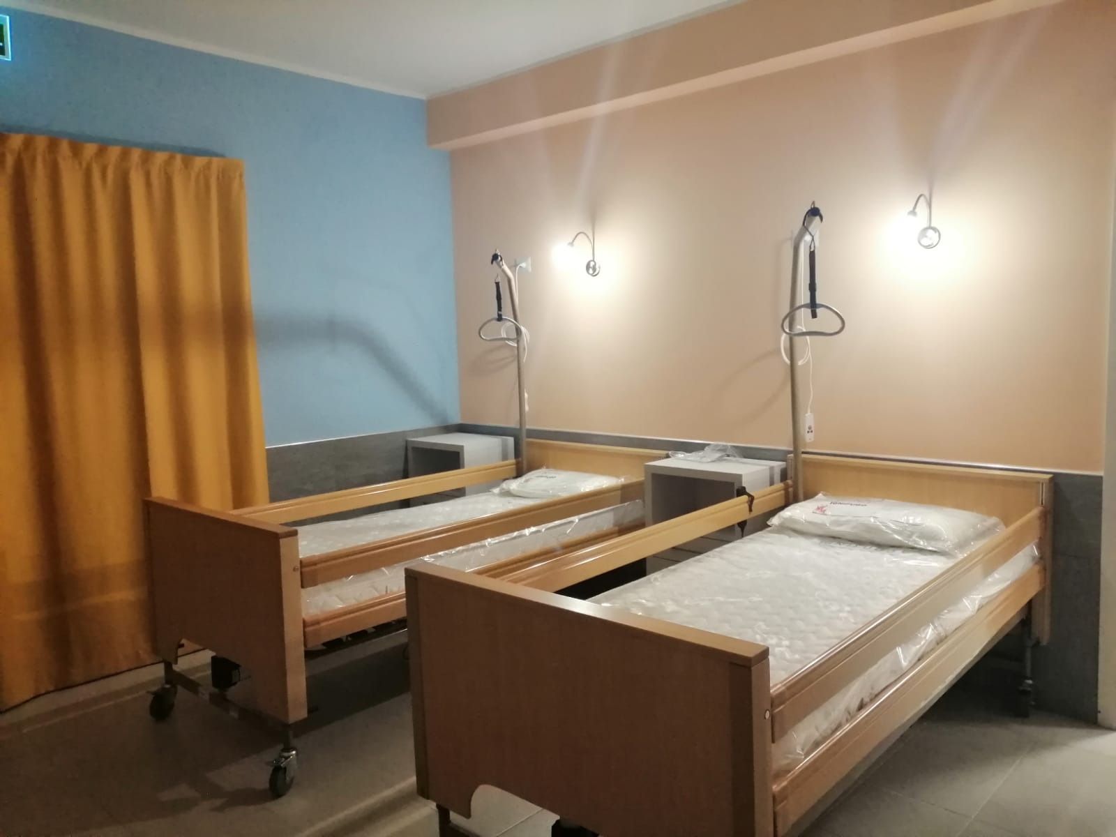 Due letti d'ospedale in una stanza con pareti color pesca chiaro e blu, tende gialle e illuminazione dall'alto.