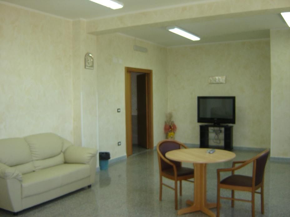 Una sala d'attesa con un divano beige, sedie, un tavolo e una TV; una porta conduce a un'altra stanza.