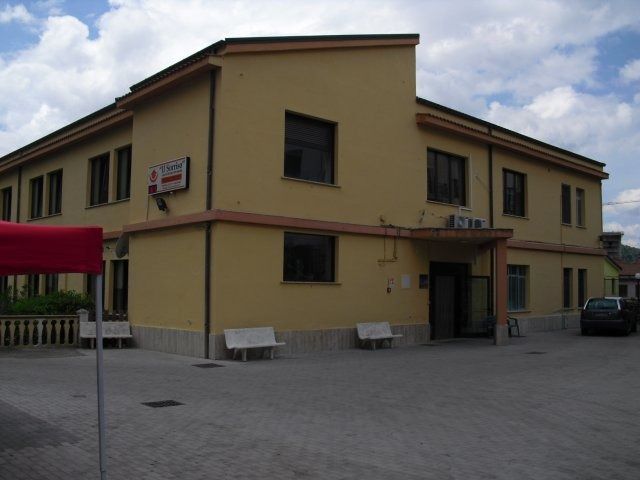 Edificio giallo a due piani con più finestre. Tre panchine all'esterno su un terreno asfaltato.