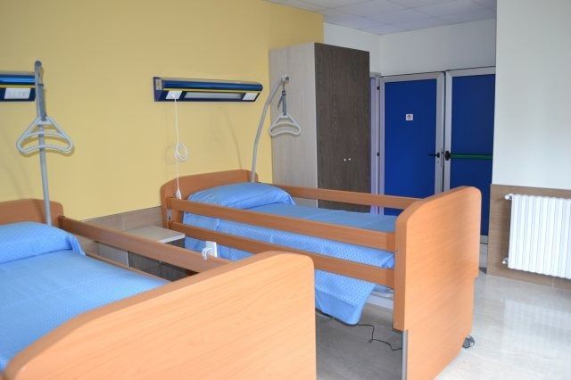 Due letti d'ospedale in una stanza con lenzuola blu, pareti gialle e porte blu.