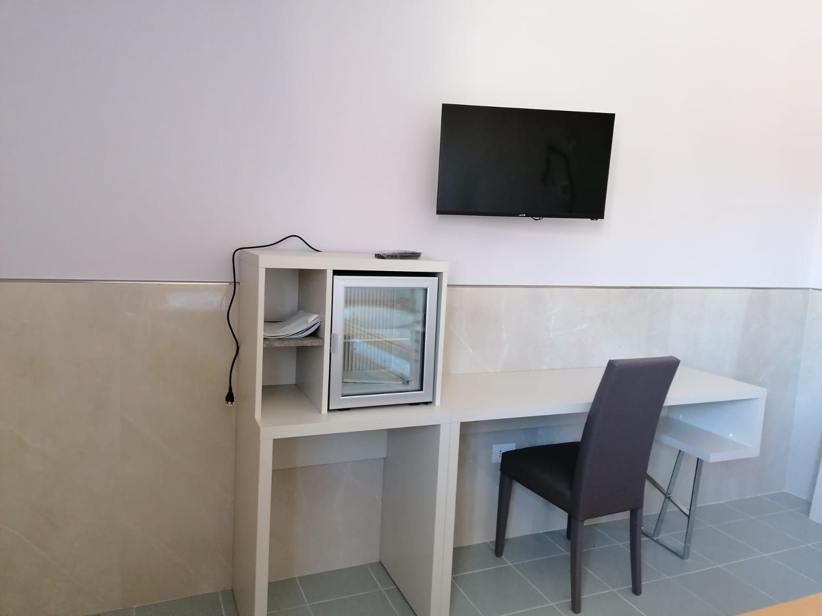 Una piccola scrivania con minifrigo e TV a parete. Una sedia è posizionata accanto alla scrivania.