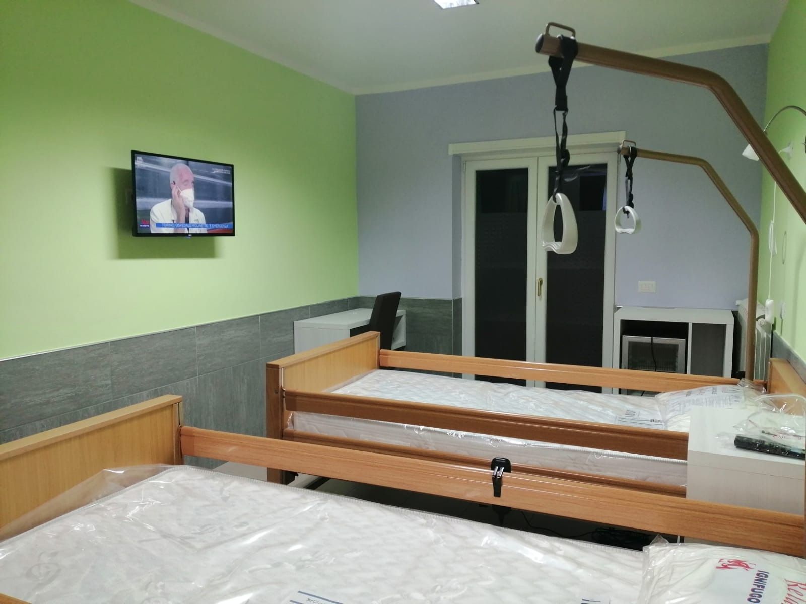 Due letti d'ospedale in una stanza con TV, ascensore e porta che si apre su un balcone. Pareti verdi e blu.