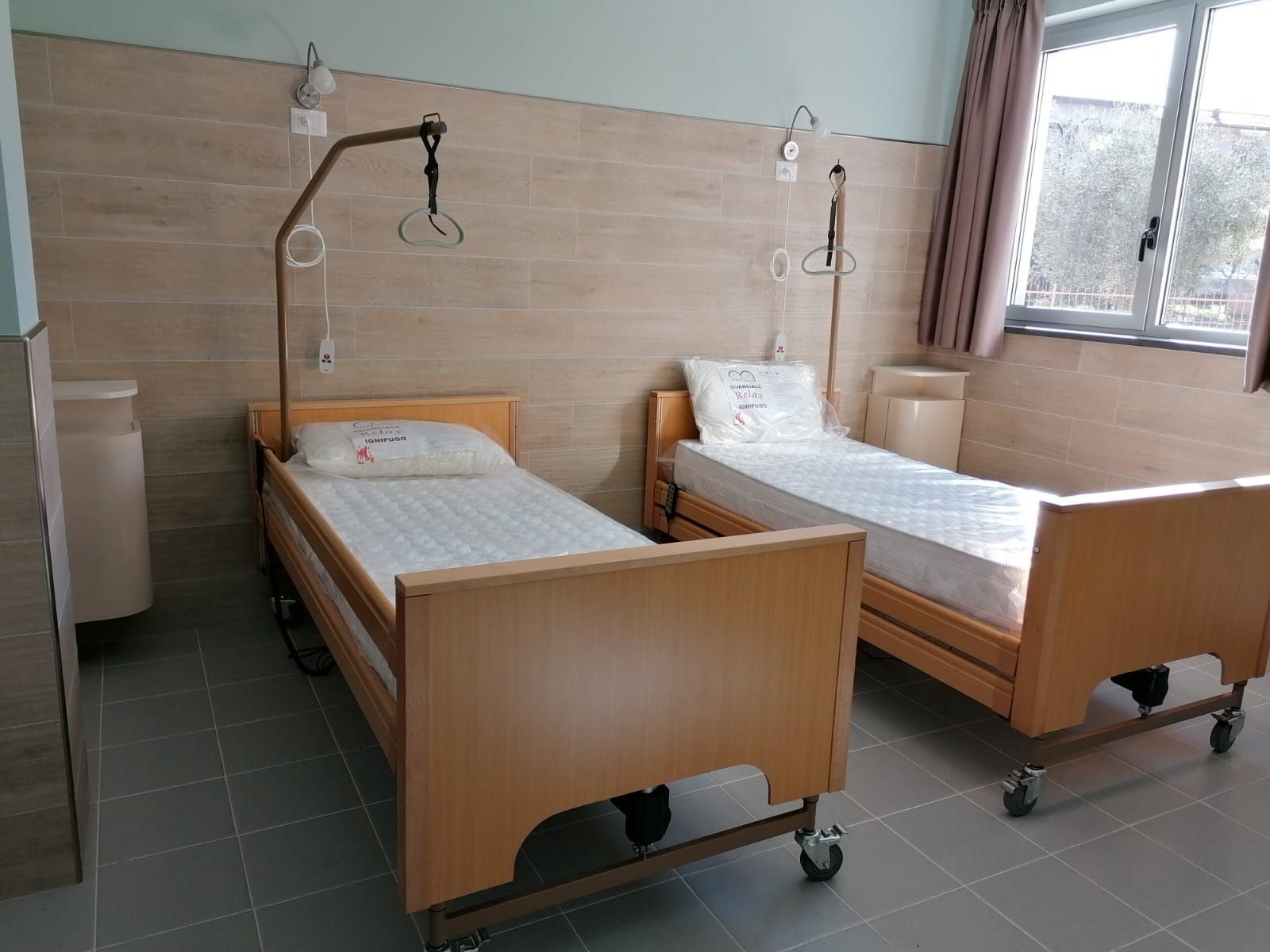 Due letti d'ospedale in una stanza, pareti e tende beige, apparecchiature mediche visibili, una finestra sullo sfondo.