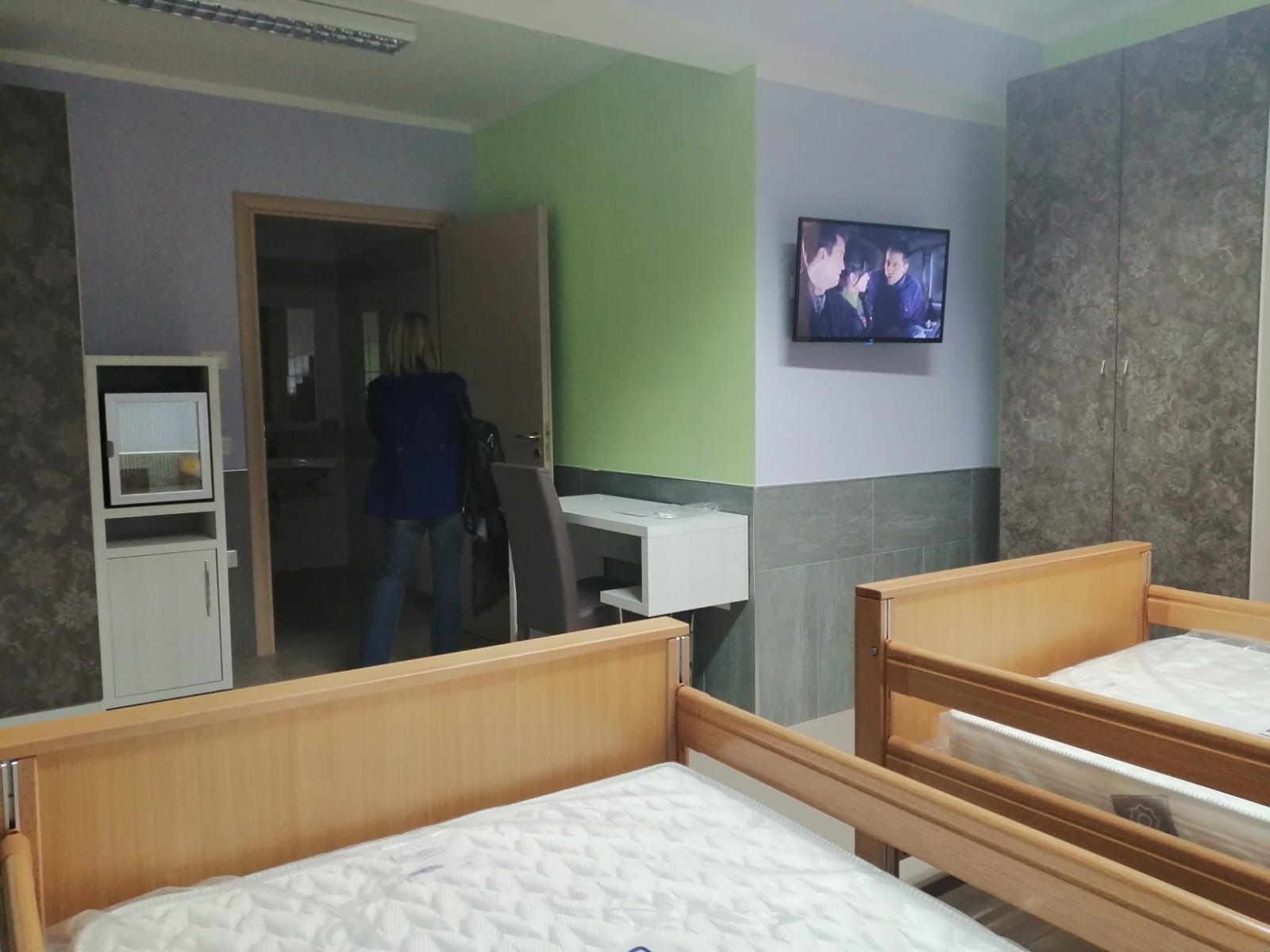 Due letti in una stanza con TV, porta aperta e persona. Legno chiaro e pareti di vari colori.