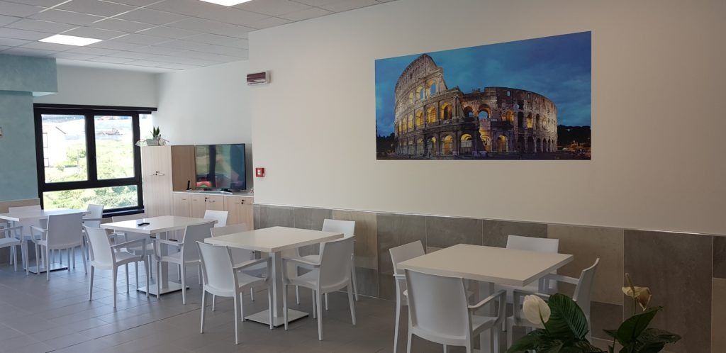 Interno di una sala da pranzo con tavoli, sedie e un'opera d'arte raffigurante il Colosseo.