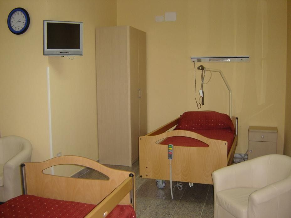 Stanza d'ospedale con due letti, sedie, TV e apparecchiature mediche. Pareti giallo pallido.