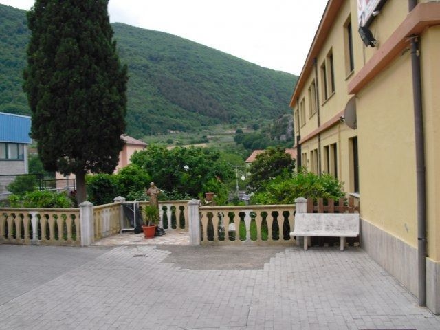Edificio con cortile, montagne sullo sfondo; sono visibili alberi e una panchina.