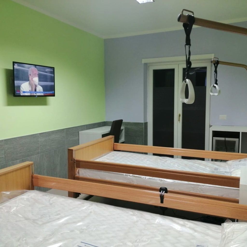 Due letti d'ospedale in una stanza con TV, attrezzature e porta. Pareti verdi e blu.