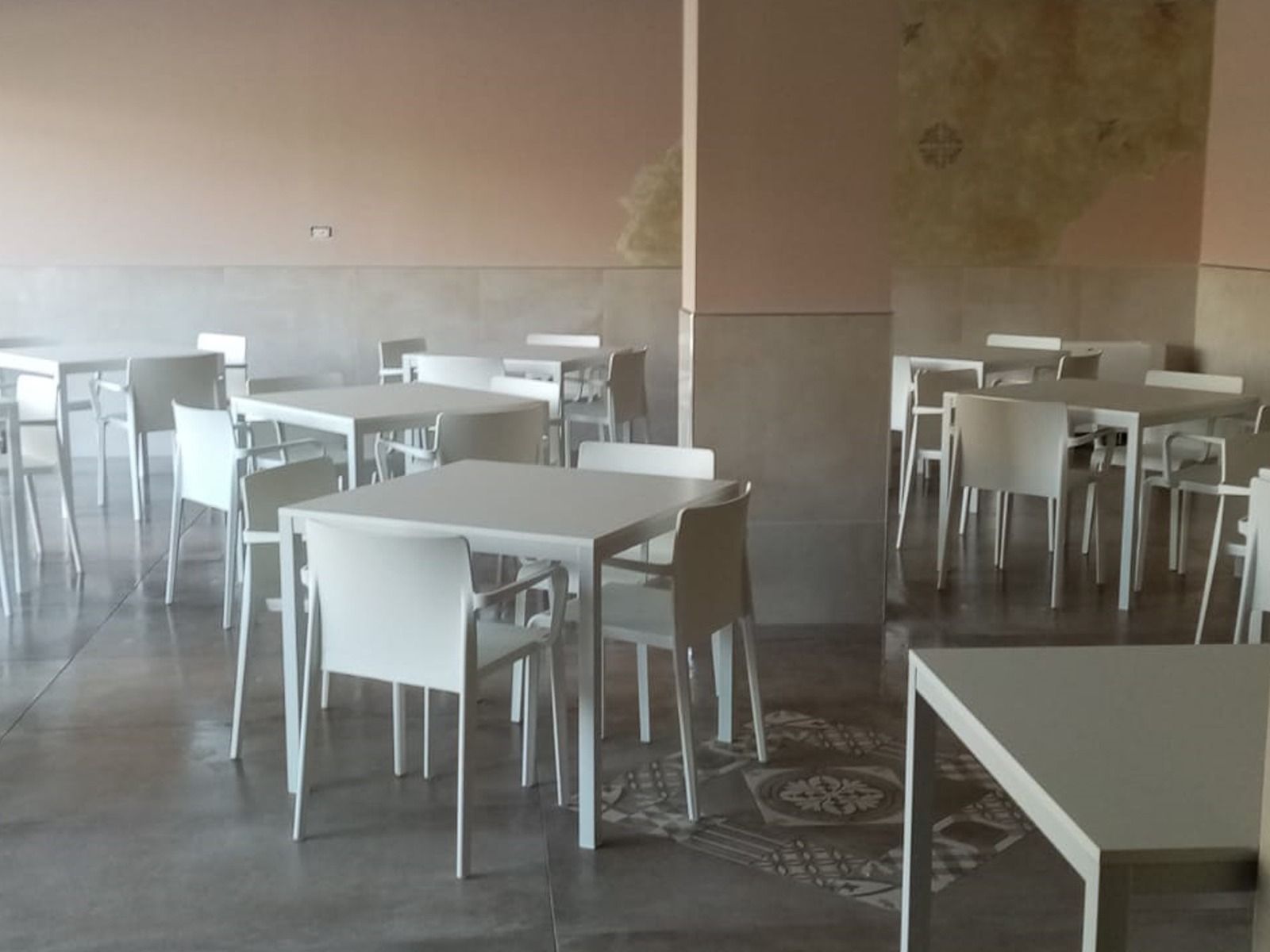 Stanza vuota con tavoli e sedie bianchi quadrati. Le pareti sono rosa pallido e beige.