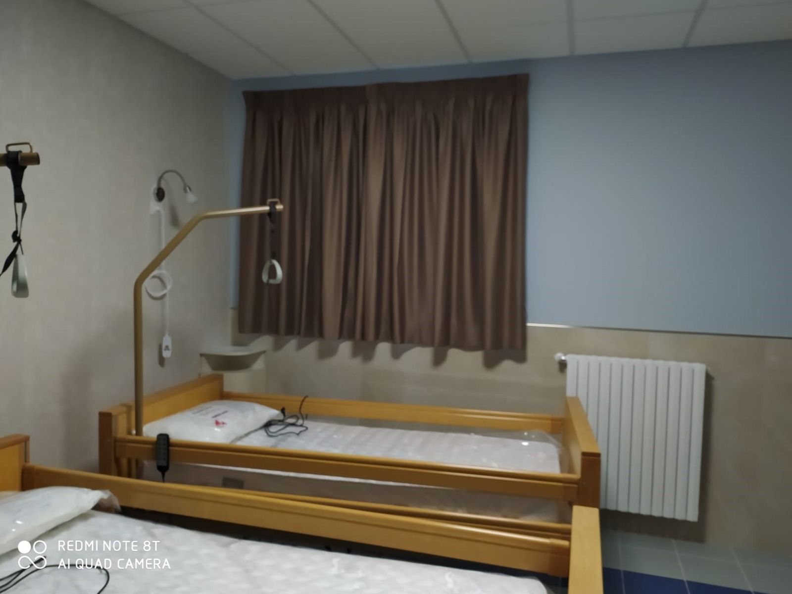 Stanza d'ospedale con due letti, finestra con tende marroni, pareti blu e beige, termosifone.