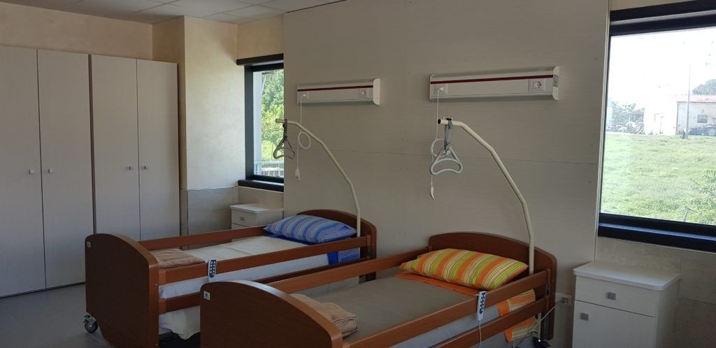 Due letti d'ospedale in una stanza con un armadio e finestre che si affacciano su un prato.