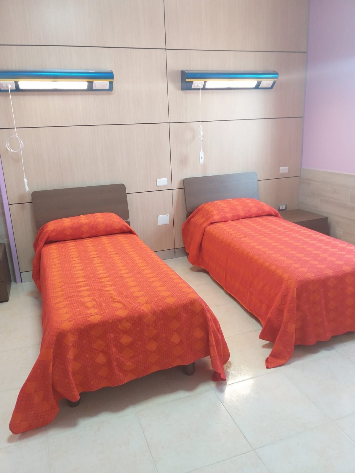 Due letti singoli con lenzuola arancioni in una stanza dai colori chiari; due luci a parete sopra.