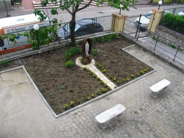 Giardino interno con statua, panchine e un autobus. Terreno marrone, sentiero pavimentato e recinzione.