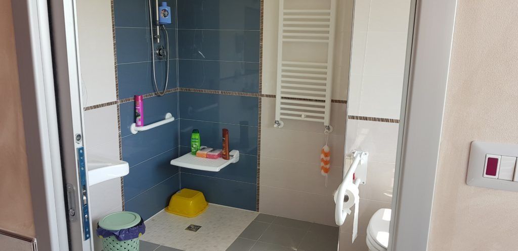 Un bagno con piastrelle blu, una sedia per la doccia e uno scaldasalviette. Un secchio giallo è appoggiato sul pavimento.