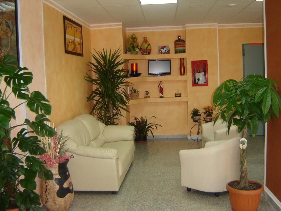 Sala d'attesa con divani bianchi, piante in vaso, TV a parete e mensole decorative.