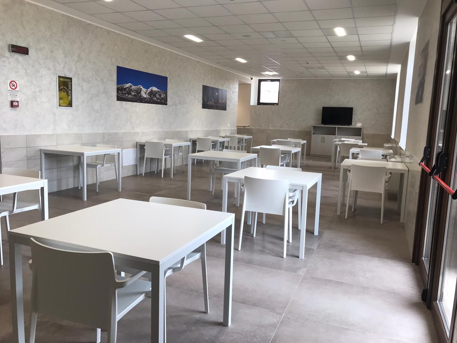 Stanza vuota con tavoli e sedie bianchi, una TV e opere d'arte paesaggistica.