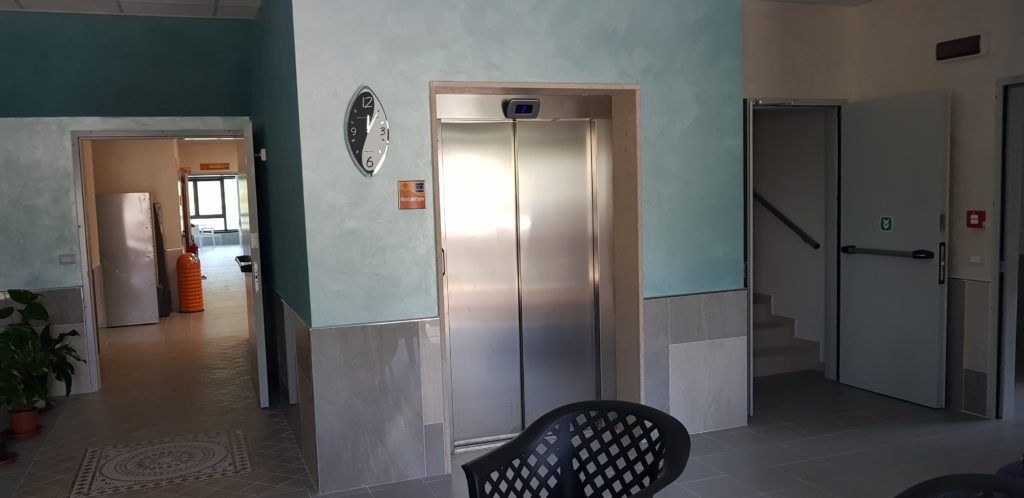 Un'inquadratura interna di un edificio con un ascensore e una porta aperta. Le pareti sono azzurre e beige.