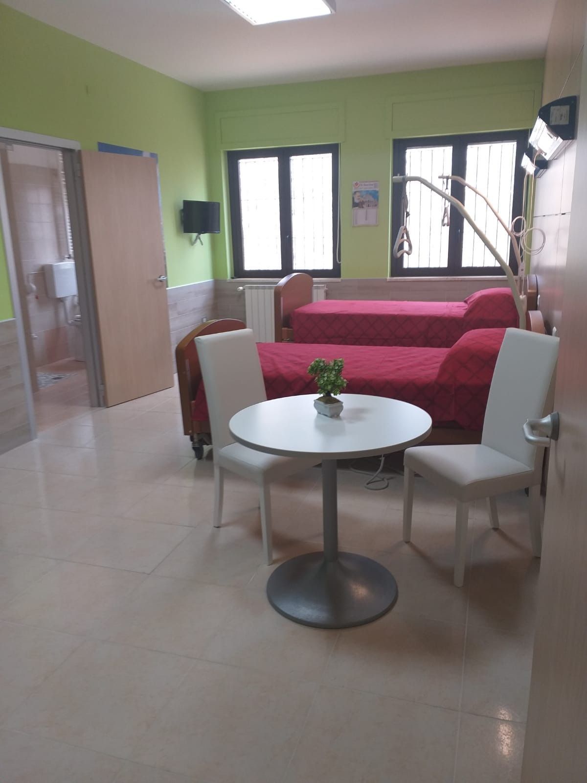 Camera con due letti, tavolino e sedie. Pareti verdi, copriletto rossi e porta del bagno.