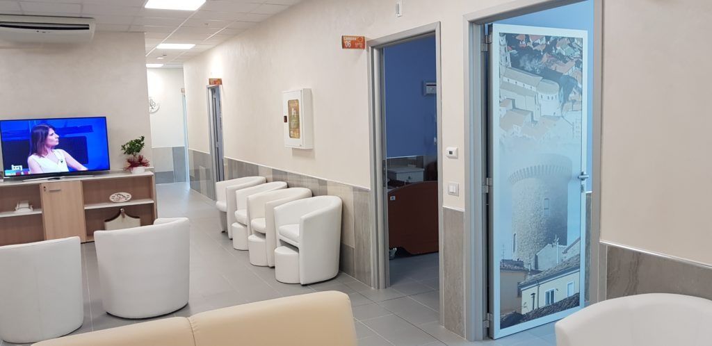 Una sala d'attesa ben illuminata con sedie bianche, una televisione e due porte aperte.