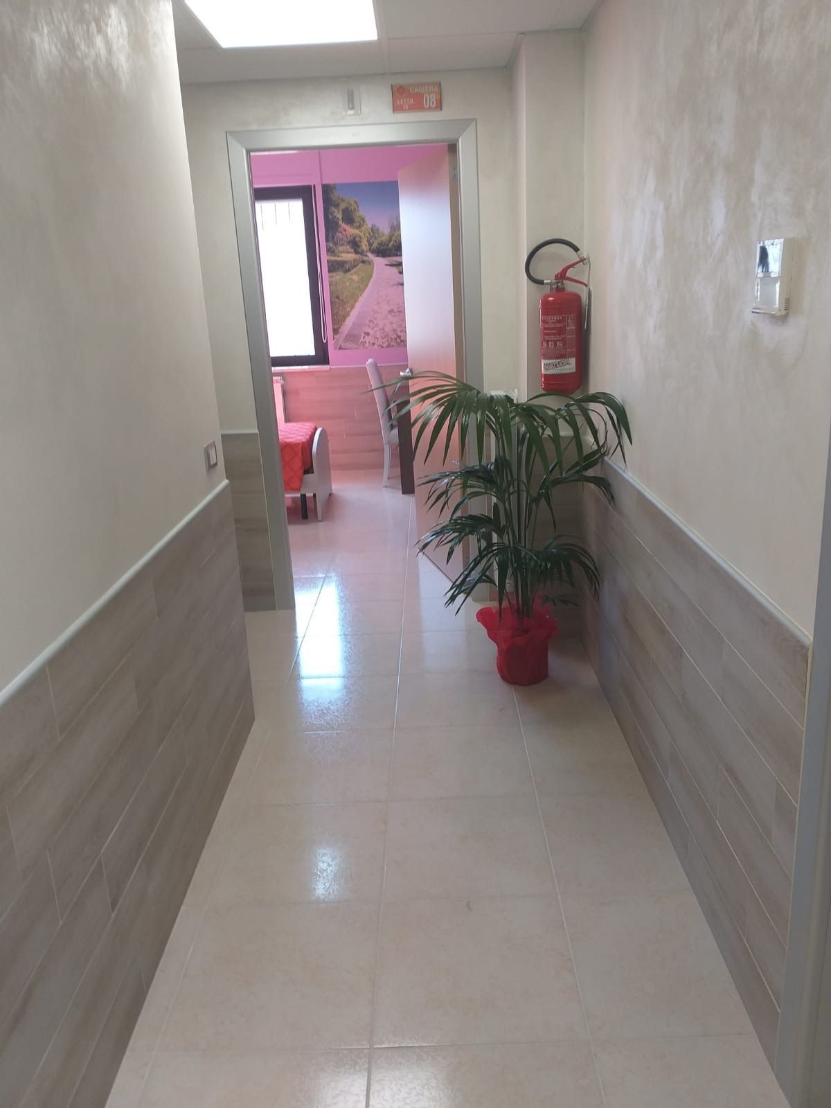Corridoio stretto con pavimento piastrellato, pianta, estintore, che conduce a una stanza con tavoli.