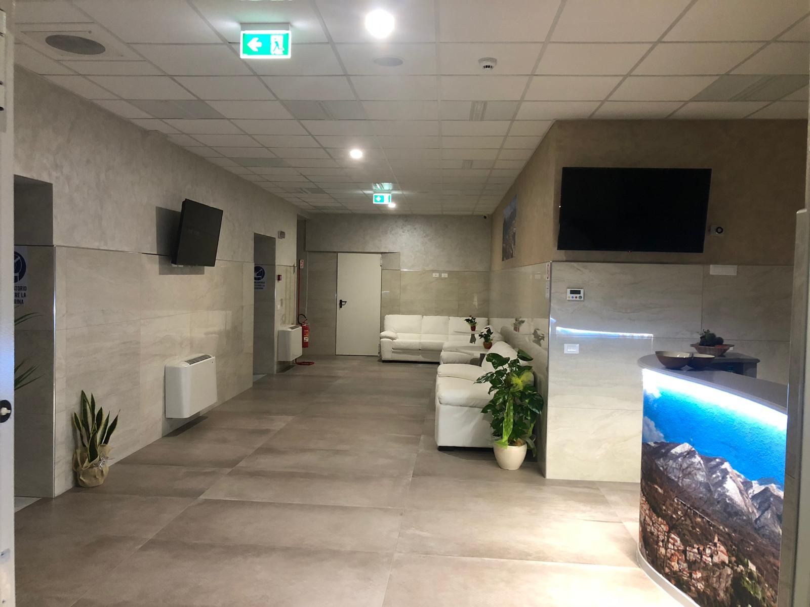 Lungo corridoio con sala d'attesa in una moderna clinica medica. Pareti e pavimento in cemento. Schermi TV, divano bianco, reception.