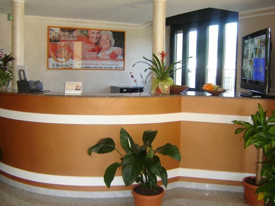 Reception con piante, poster e TV in un edificio. Colori marrone, bianco e beige.