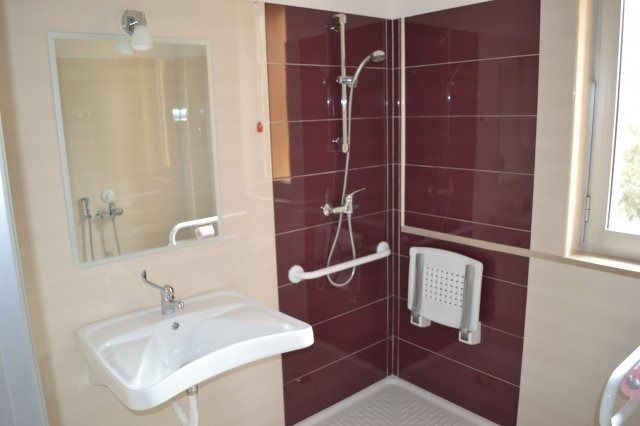 Bagno accessibile con doccia, lavandino e corrimano. Doccia con piastrelle rosse, sedia bianca e pareti beige.