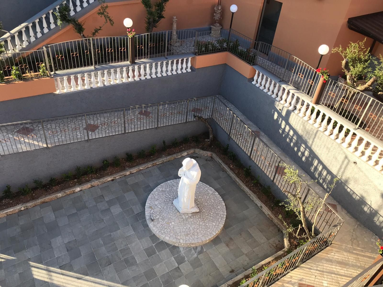 Cortile con statua al centro, circondato da ringhiere e gradini, con vista elevata.