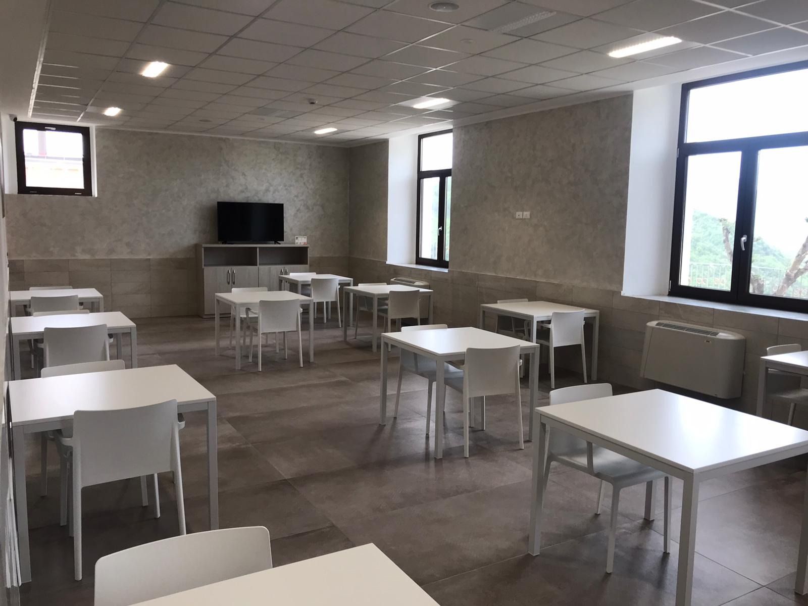 Stanza vuota con tavoli e sedie bianchi, finestre e un televisore su un mobile.