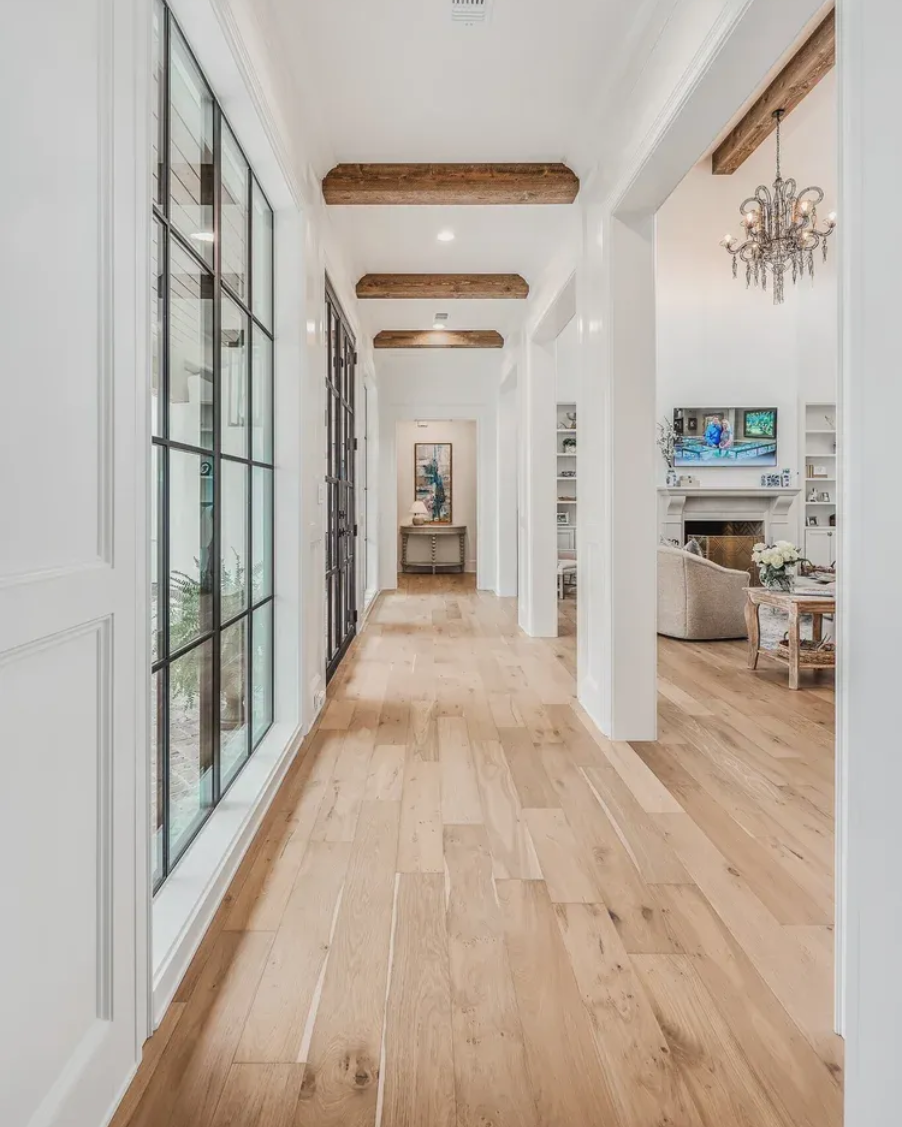 Long hallway with light wood floors, white walls, and black framed windows. Brown wooden beams and a chandelier are visible.