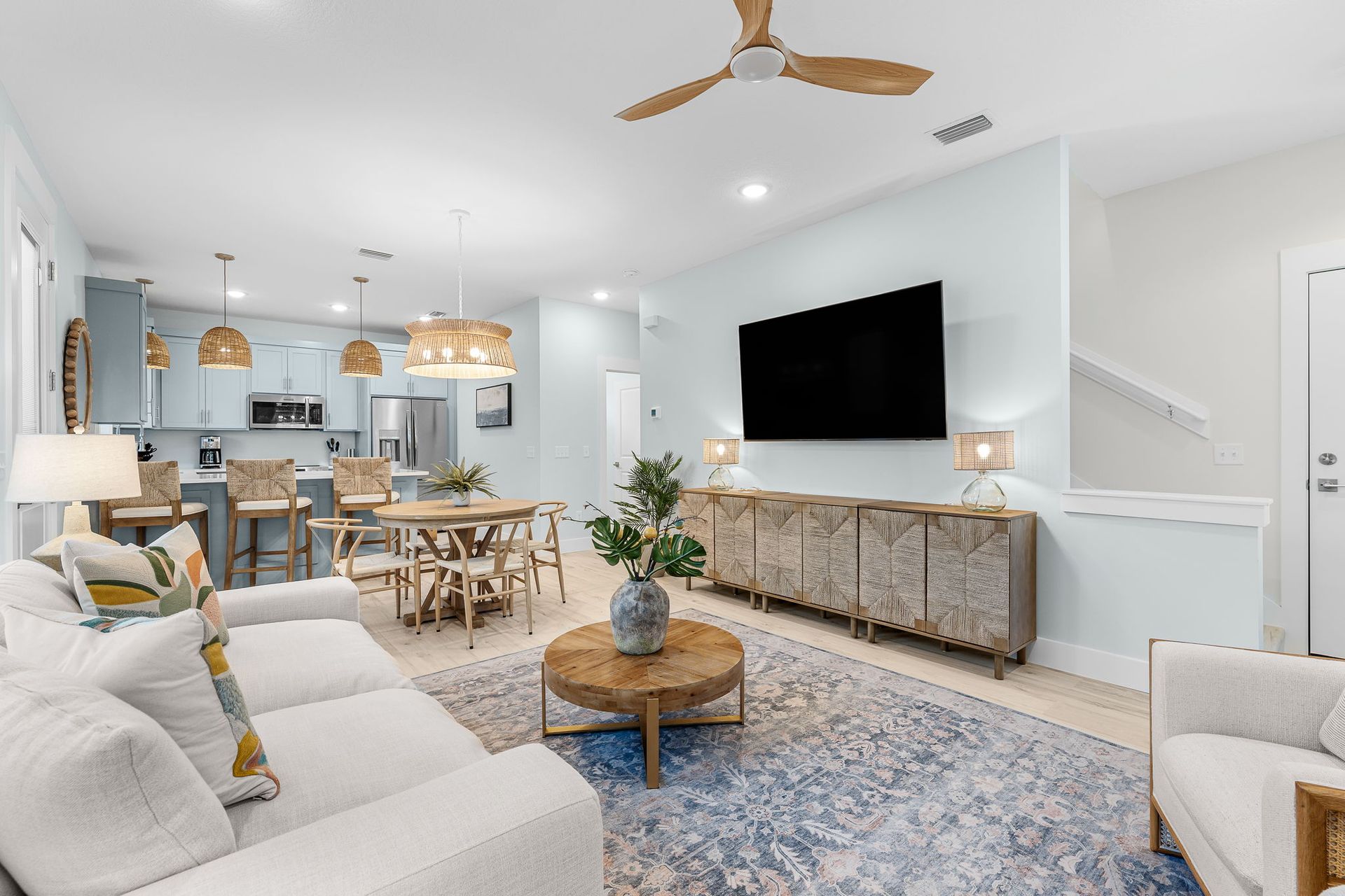 Bright modern living room with white sofas, blue rug, wall-mounted TV, and dining area in the background