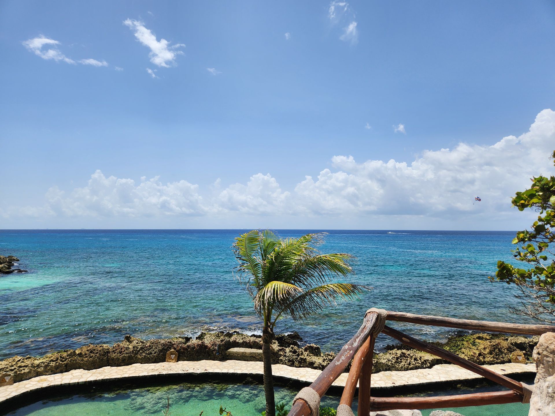 A scenic ocean view from a stone-walled overlook with a palm tree, clear blue water, and a bright, partly cloudy sky.