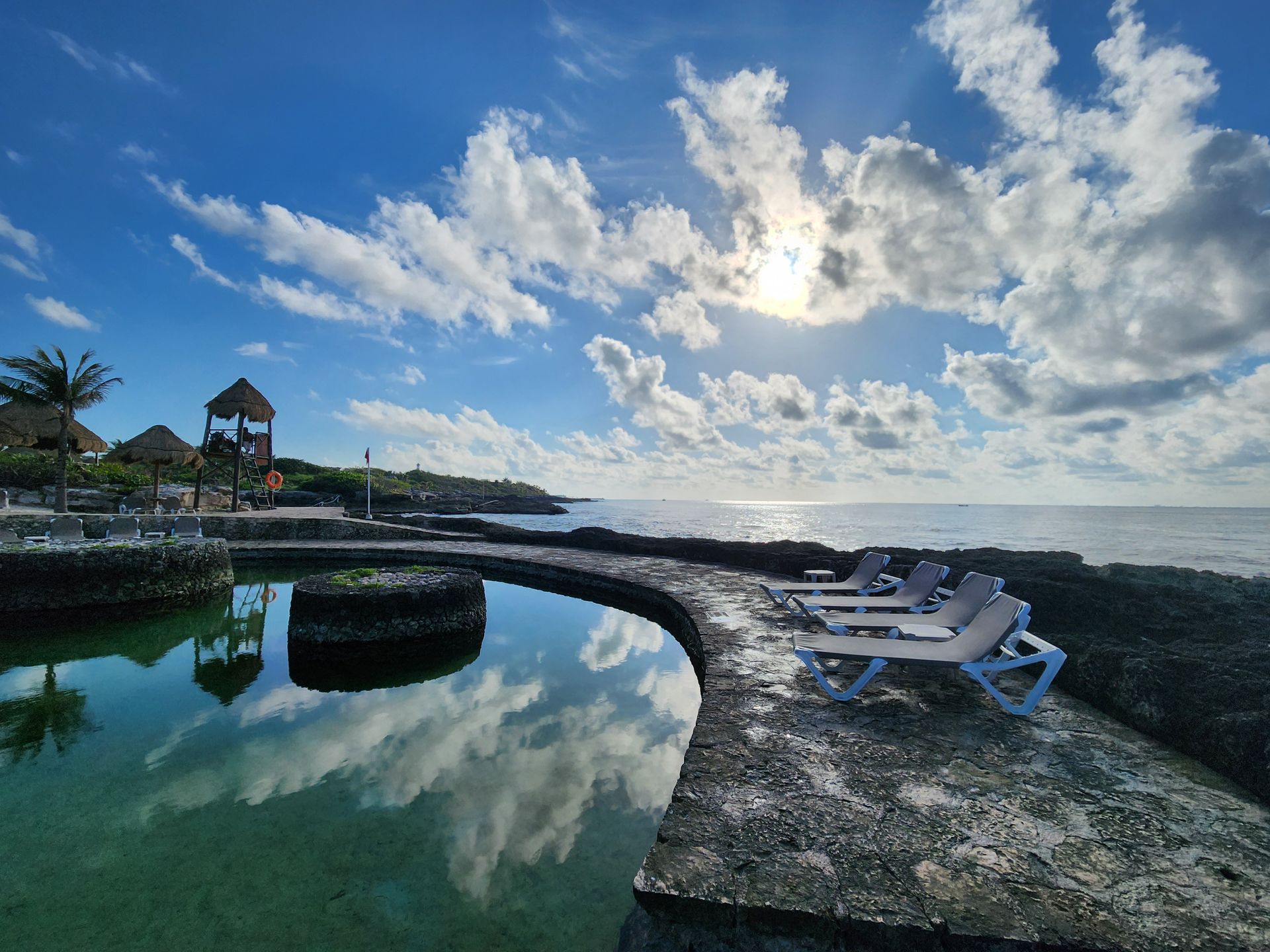 A calm, stone-rimmed lagoon by the ocean with lounge chairs on a rock patio under a bright, cloudy blue sky.