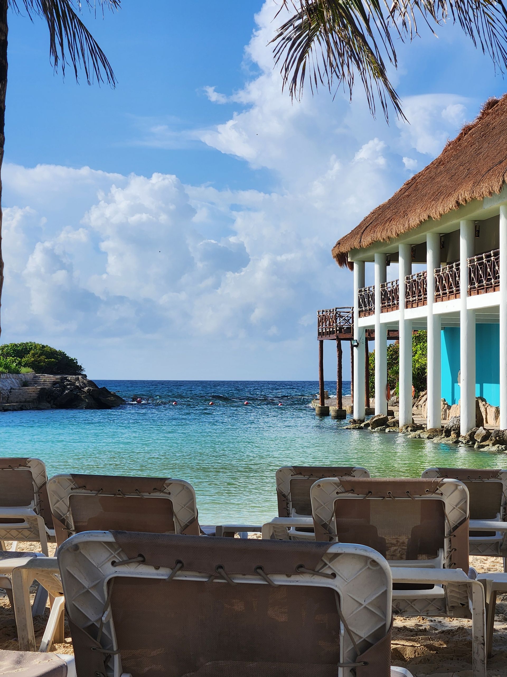 A tropical beach scene with lounge chairs in the foreground, facing a turquoise sea and a thatched-roof building on stilts.