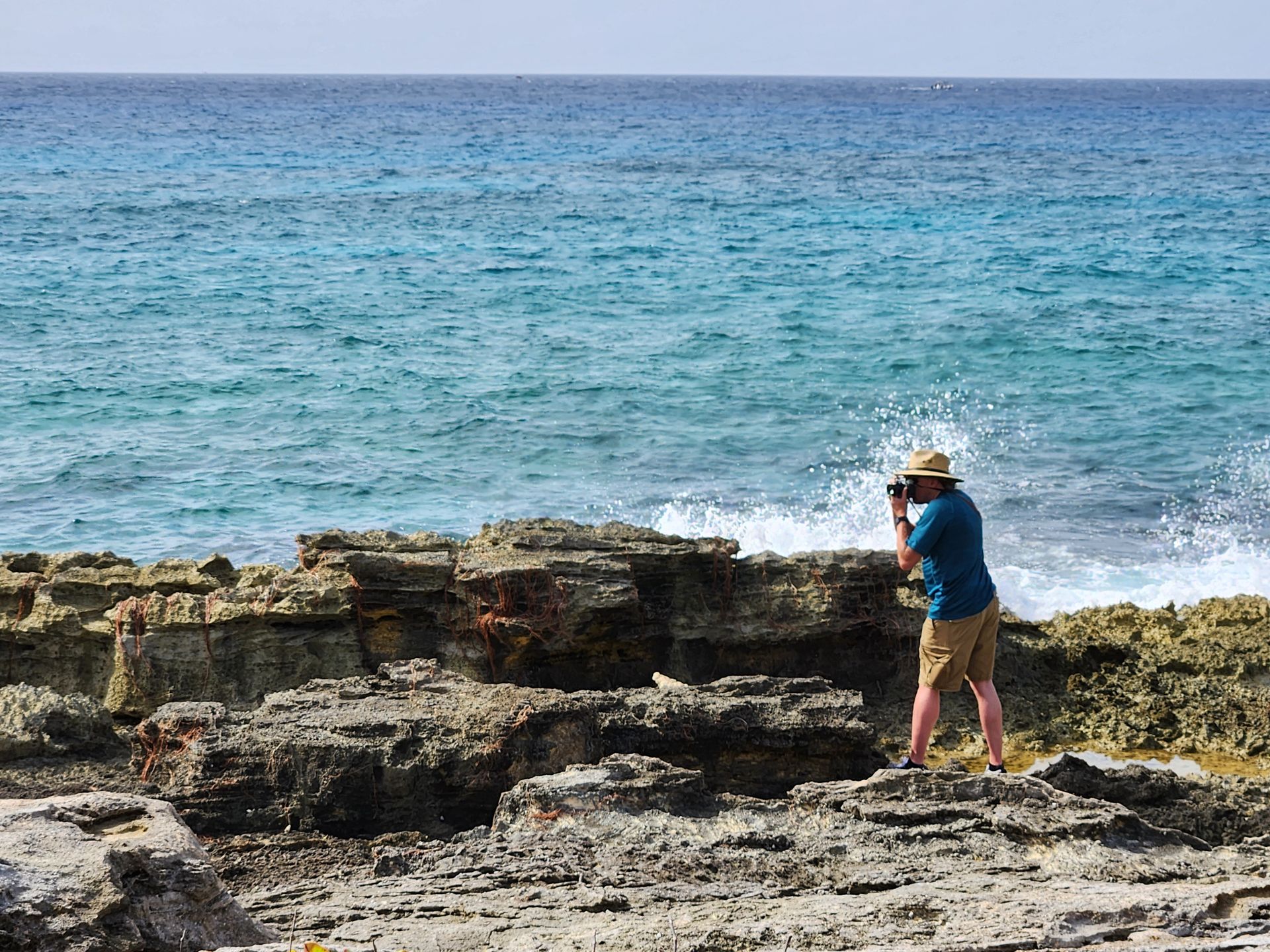 A person in a sun hat and blue shirt takes a photo of waves crashing against rocky shoreline.
