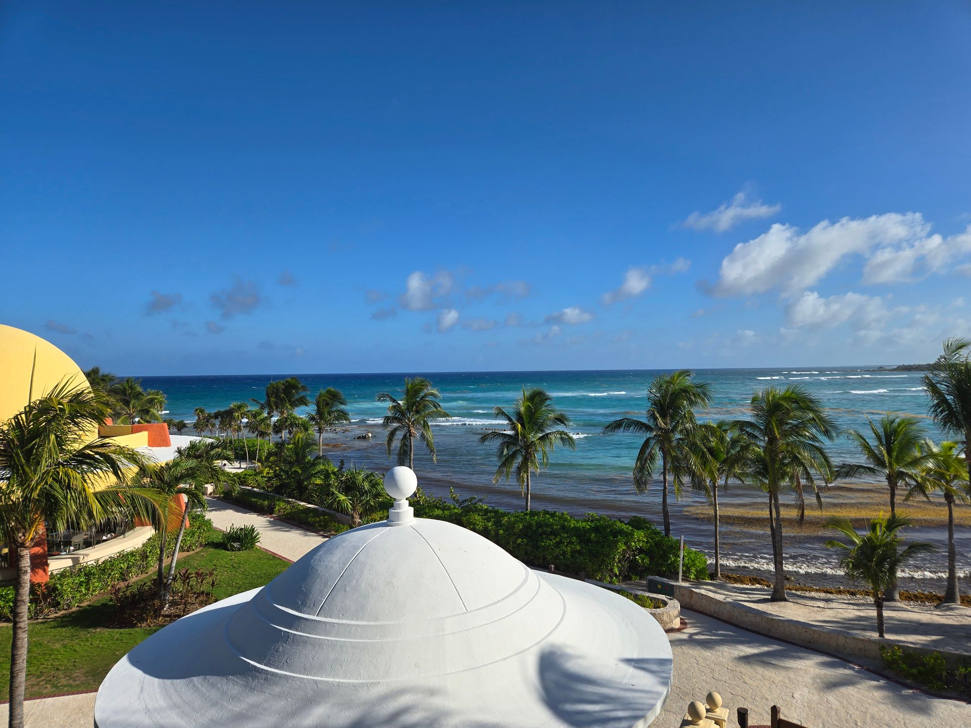 A white dome overlooking palm trees and a bright blue ocean under a clear, sunny sky.