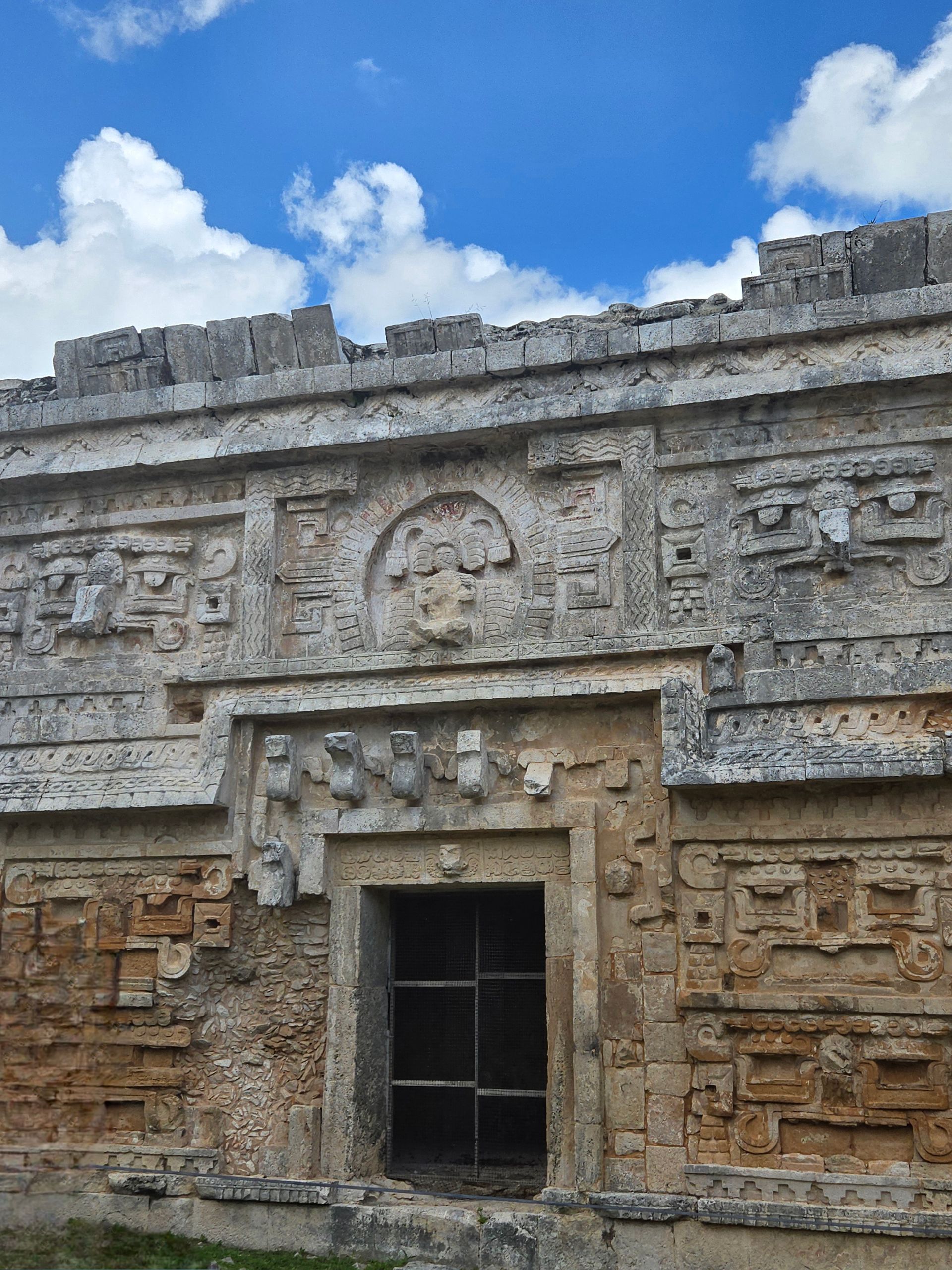 A weathered stone Mayan temple facade with intricate carvings, a central doorway, and a bright blue cloudy sky background.